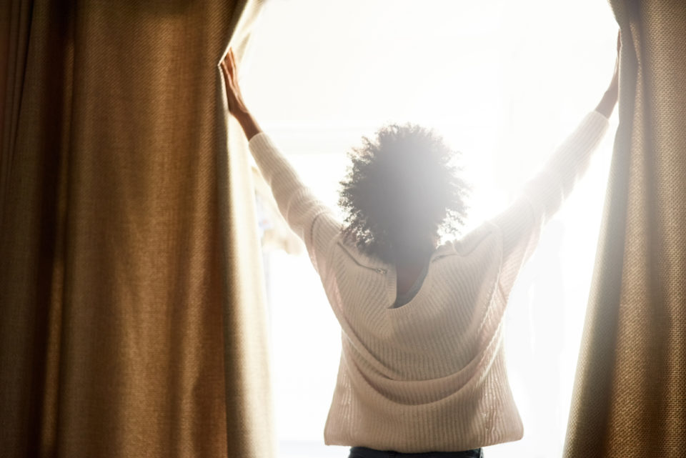 Rearview shot of a woman opening the curtains on a bright sunny day