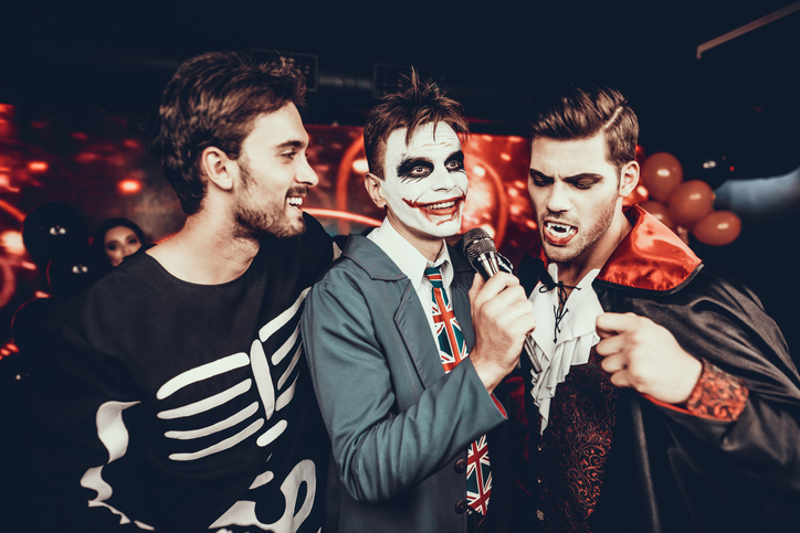 Young Men in Halloween Costumes Singing Karaoke. Happy Smiling Friends Wearing Costumes having Fun by Singing with Microphone at Halloween Party in Nightclub. Celebration of Halloween
