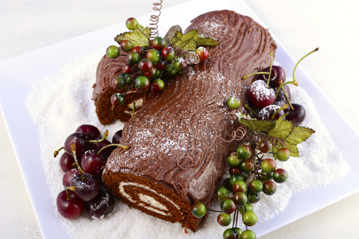 Christmas Yule Log, Buche de Noel, chocolate cake with branch, fresh cherries and festive berry decorations on a white serving platter, overhead.